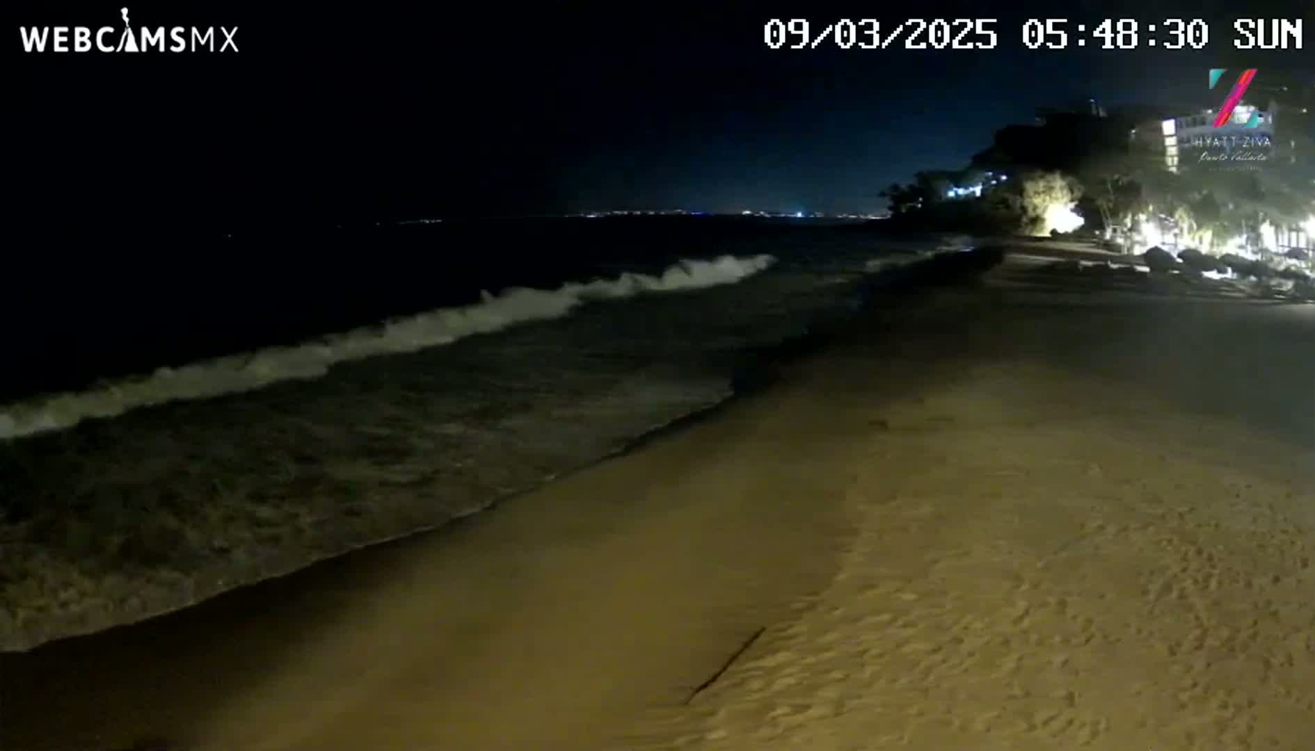 At night, moderate waves lap a sandy beach near a dimly lit building.
