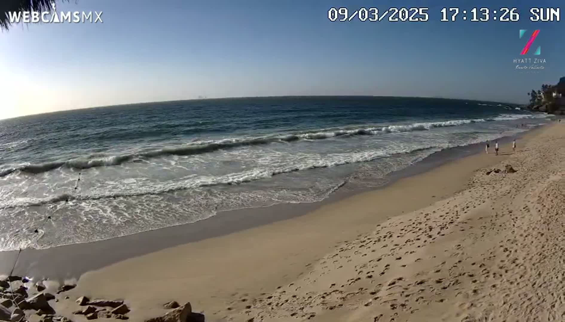 Under a clear blue sky, ocean waves roll onto a sandy beach where a few people walk.