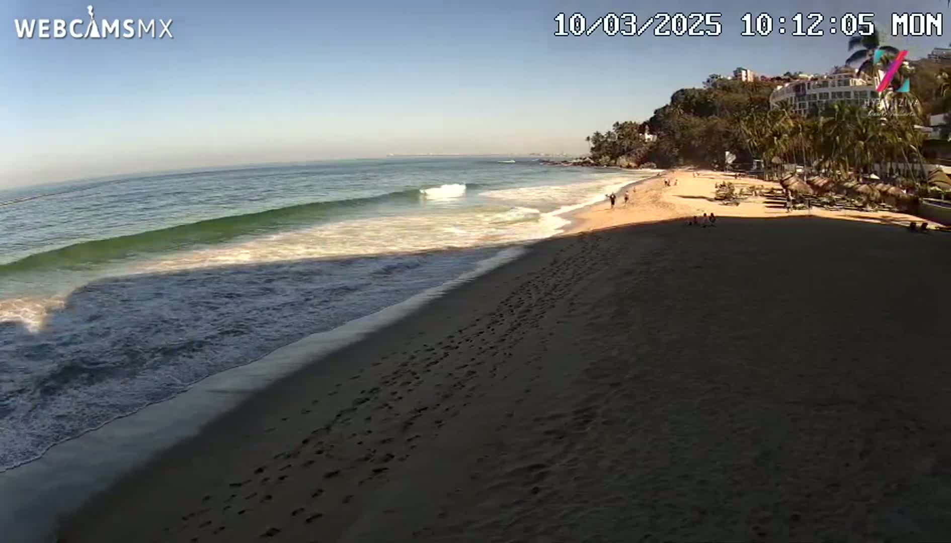 A sandy beach with footprints leading to the ocean under a clear, sunny sky shows gentle waves lapping the shore near a resort area with palm trees.