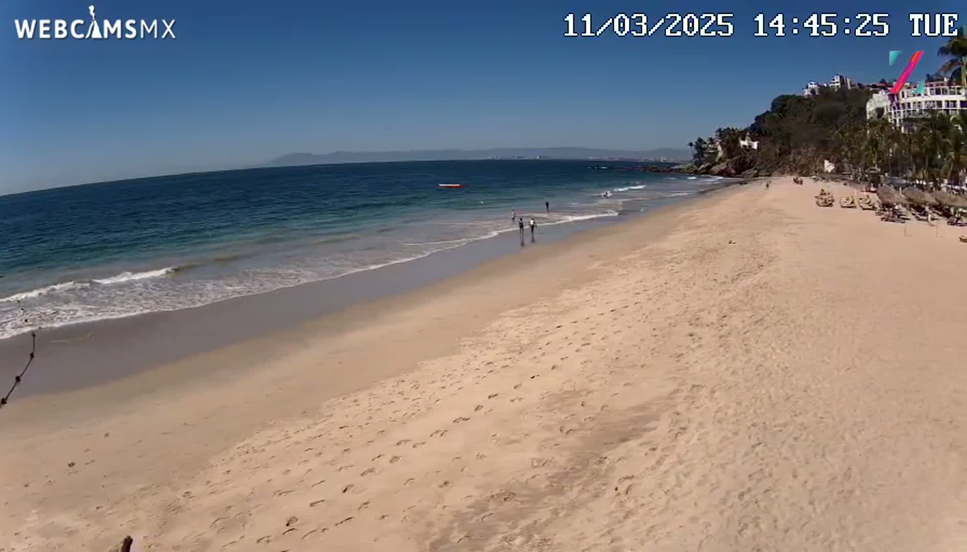 A long sandy beach with calm ocean waves under a clear blue sky, sparsely populated with people and beach chairs.