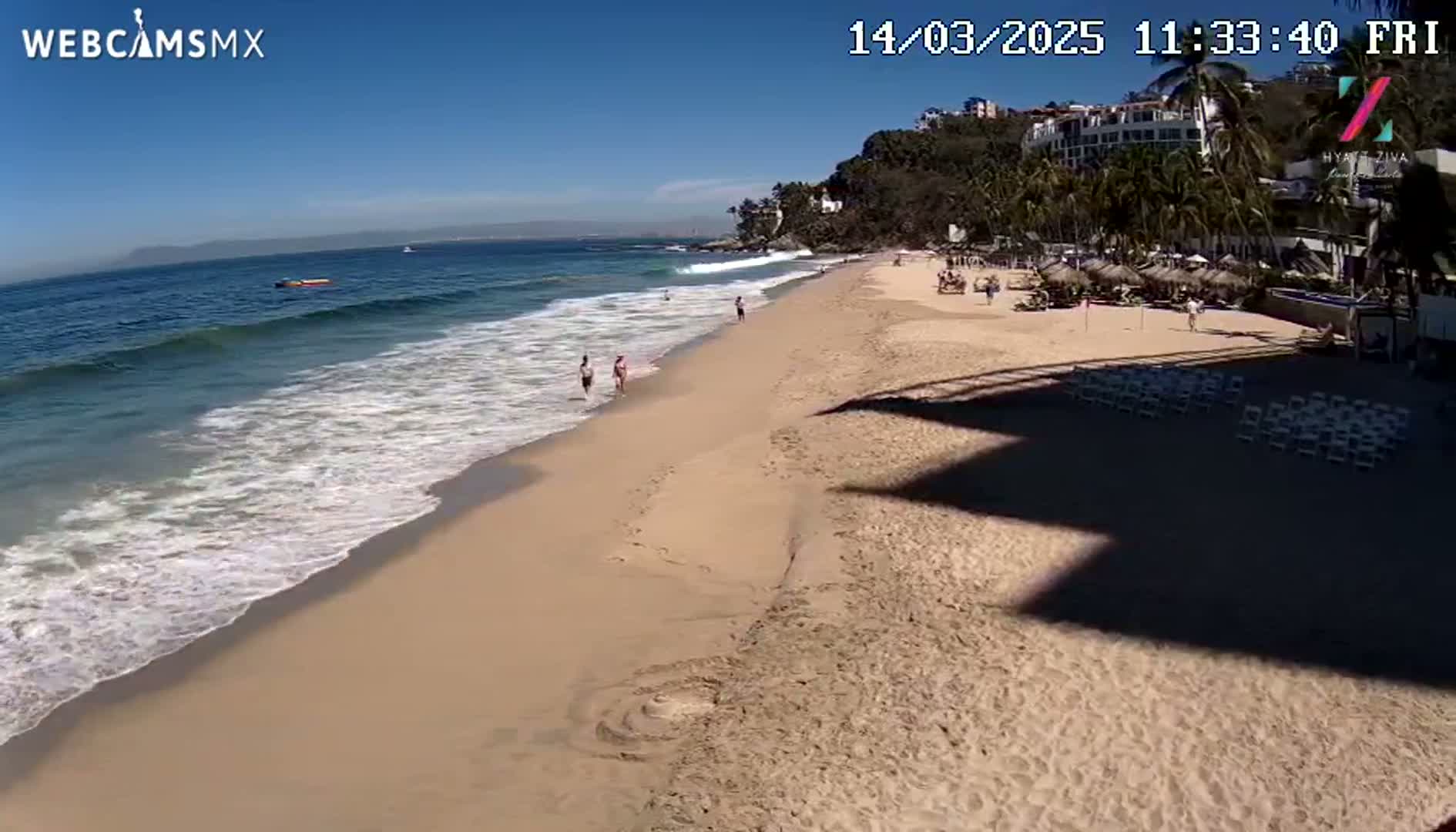 A sandy beach with gentle waves under a clear blue sky features a few people strolling along the shore and some beach umbrellas in the distance.