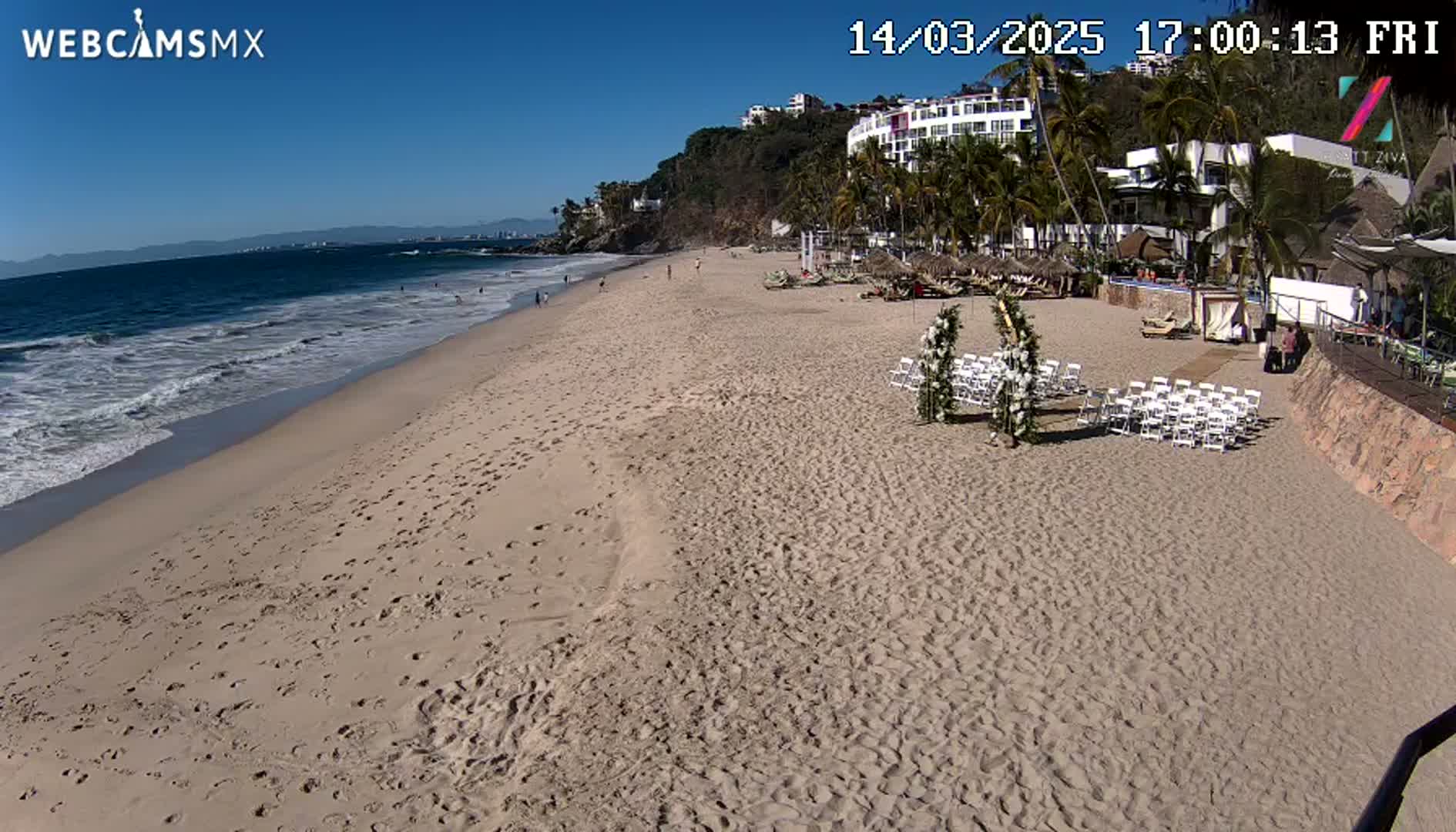 A sandy beach with a wedding setup is visible, under sunny skies, next to a calm ocean with small waves.