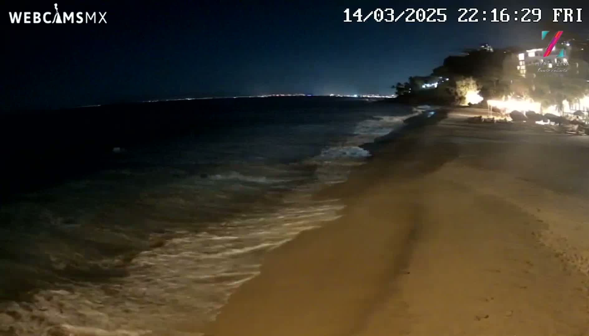 Gentle waves lap a sandy beach at night under a dark sky, with lights visible in the distance across the water and along the shoreline.