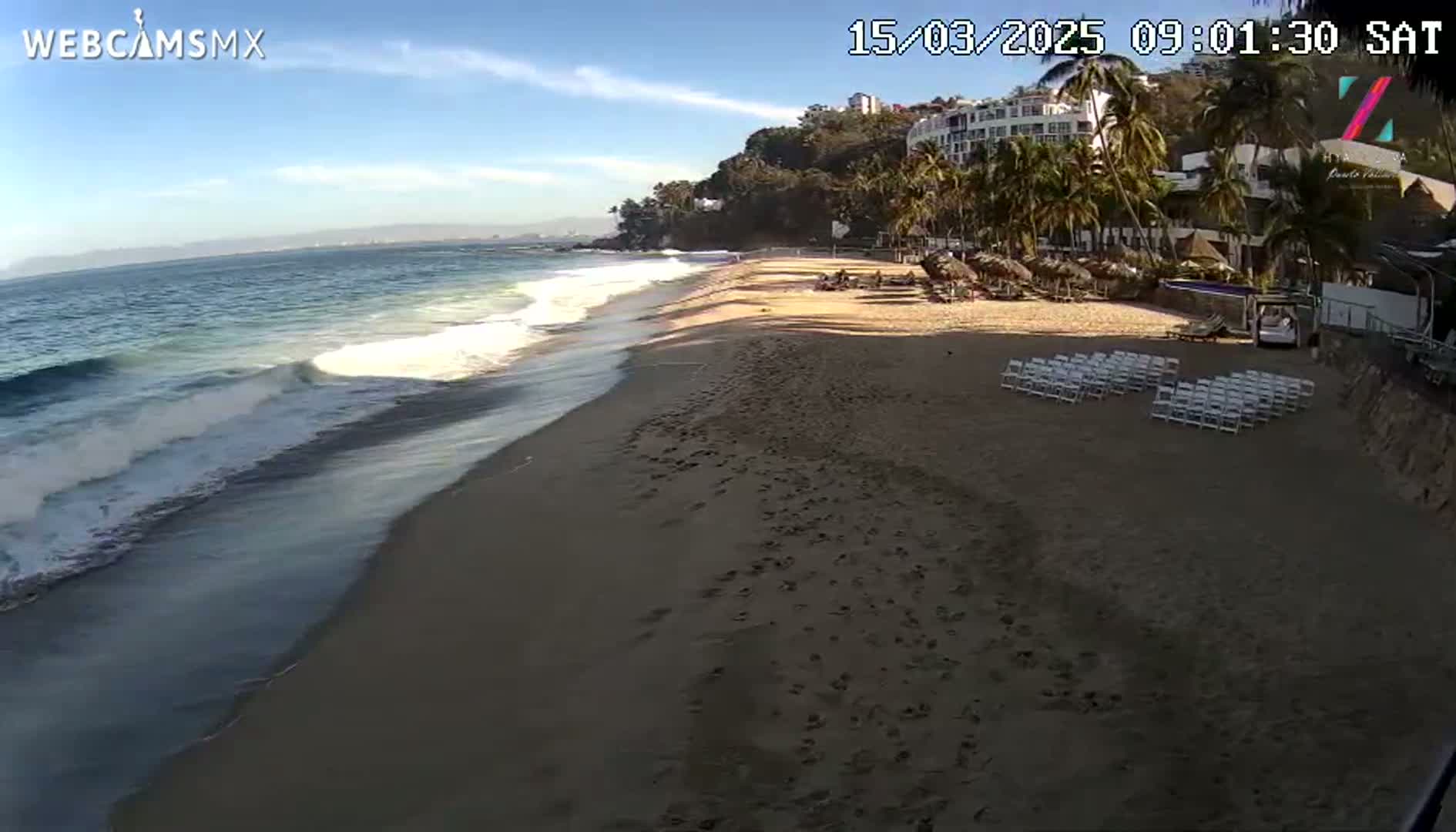 A sandy beach with gentle waves under a clear, sunny sky has rows of white chairs set up near a beachfront resort.