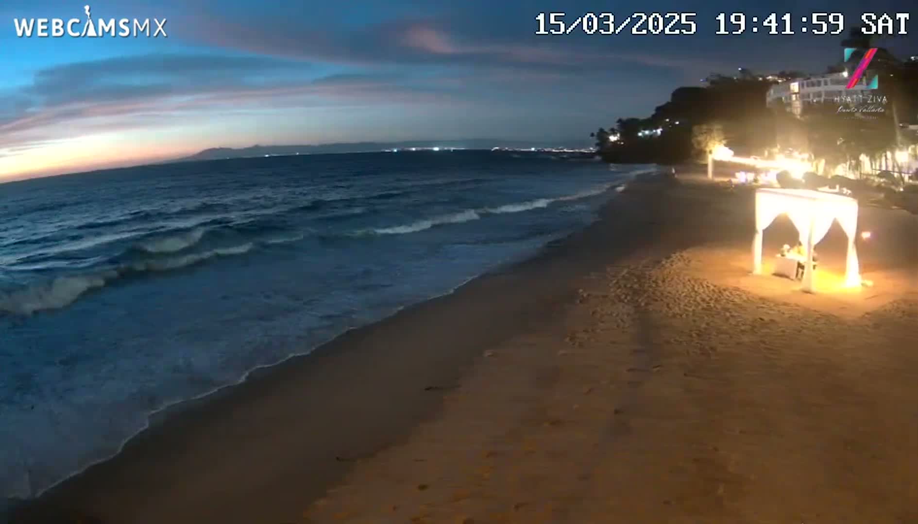 A beach at twilight shows gentle waves lapping the shore near a romantic, illuminated table set under a canopy.