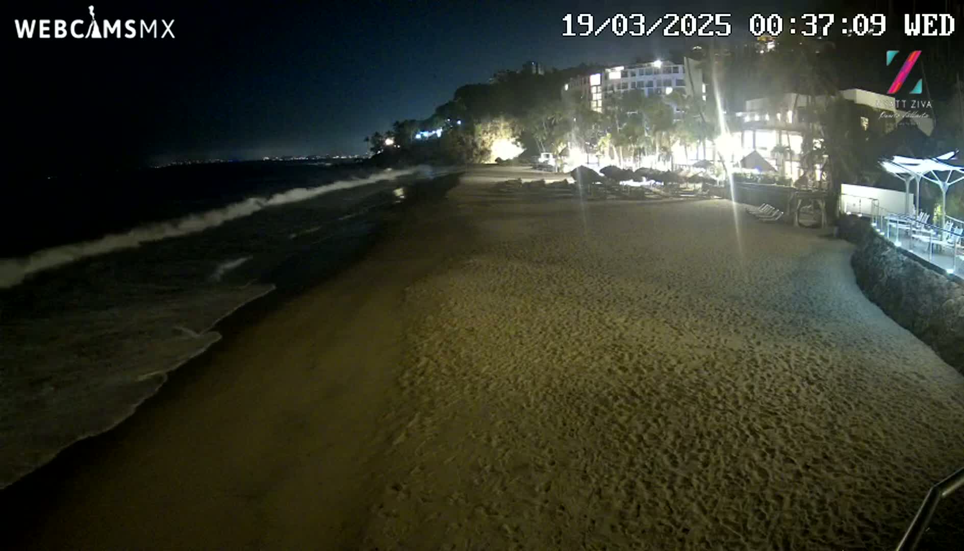 At night, gently lapping waves roll onto a sandy beach in front of a brightly lit resort nestled against a dark hillside.