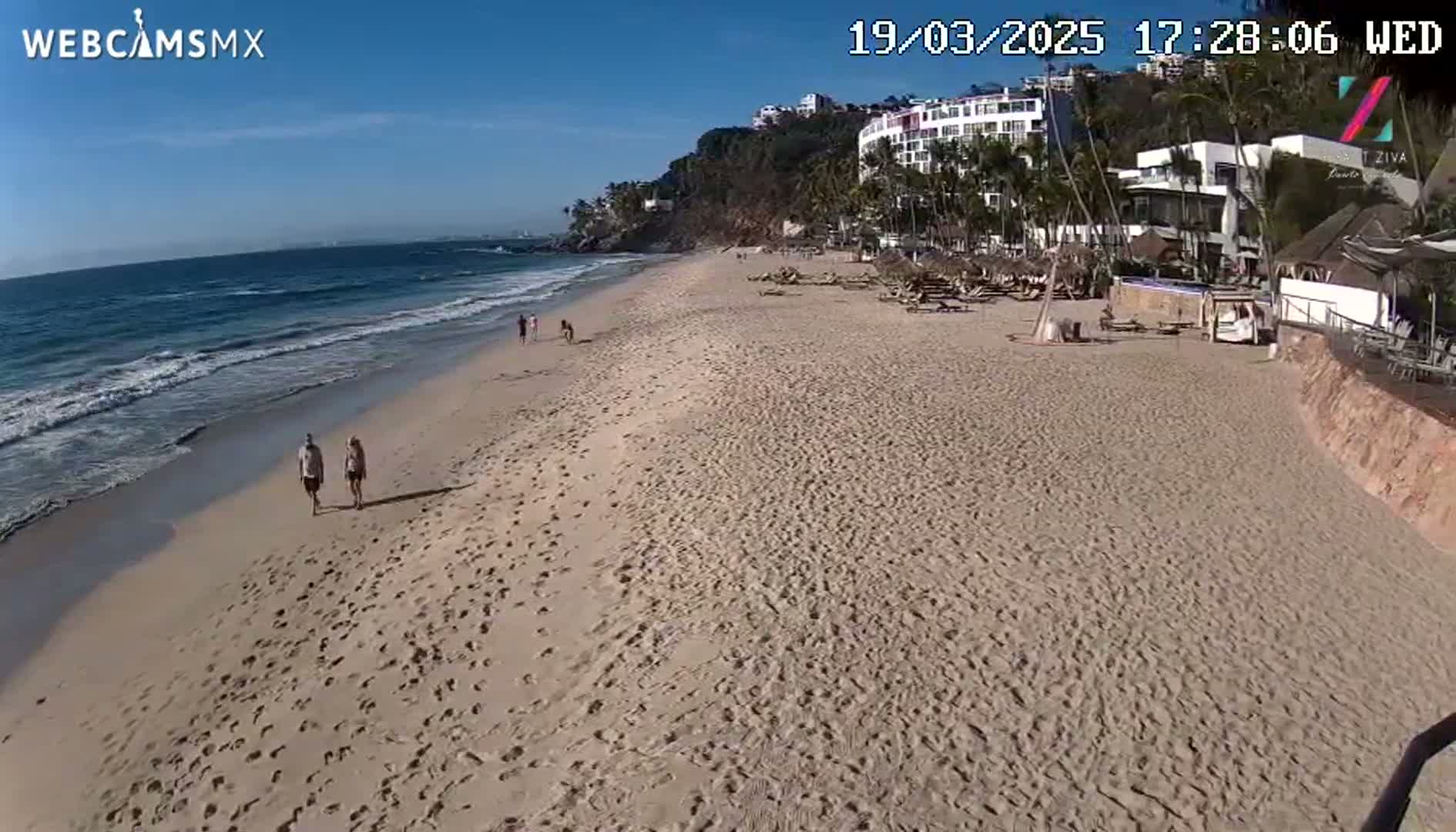 A sandy beach with a few people walking stretches from a calm ocean to buildings on a hillside under a clear blue sky.