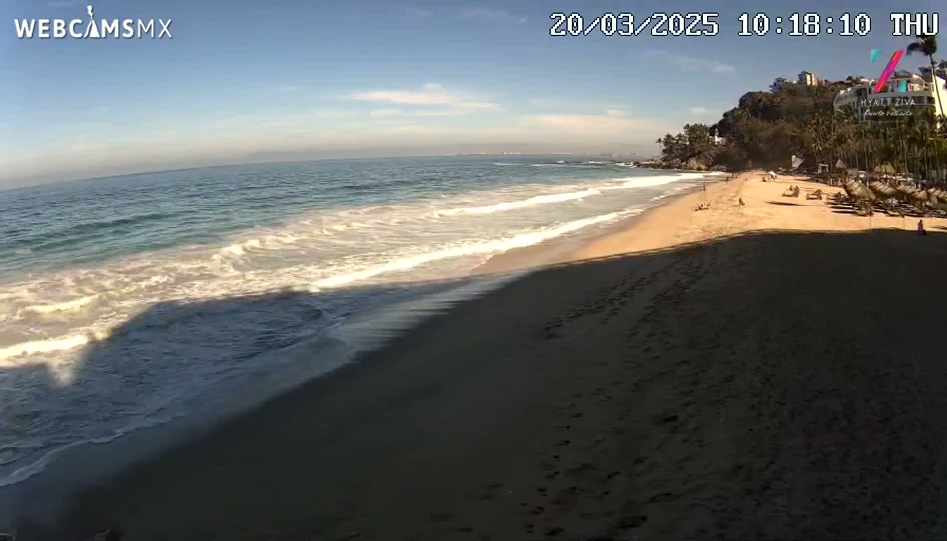 A sandy beach with gentle waves under a mostly clear sky is seen from a high vantage point.