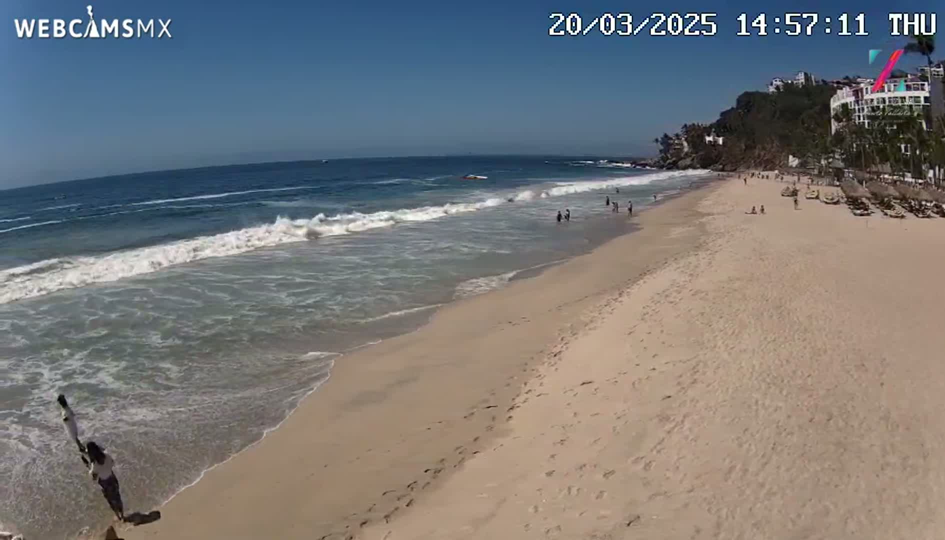 A sandy beach with gentle waves under a clear blue sky, showing a few people on the beach and in the ocean.