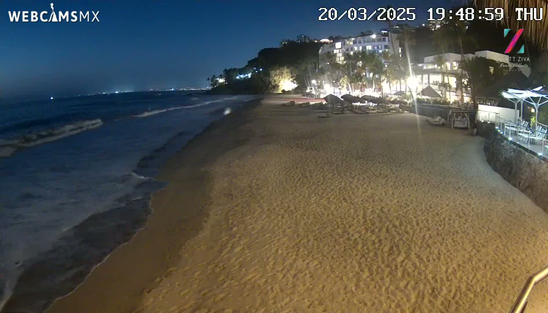 A sandy beach at night with gentle waves lapping the shore, under a dark blue sky, is backed by illuminated buildings and palm trees.