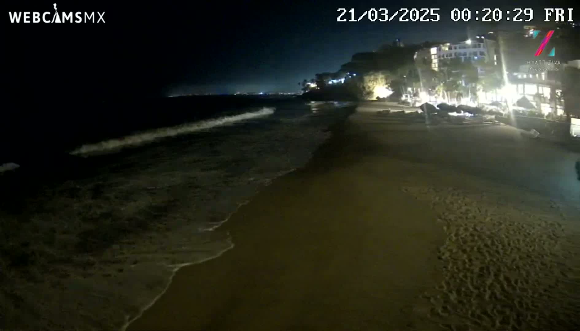 At night, gentle waves lap a sandy beach near a brightly lit resort situated on a dark cliff.