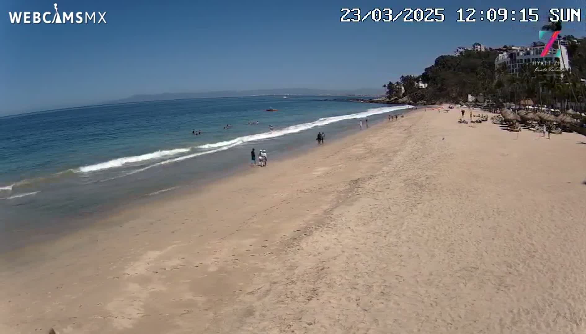 A sandy beach with a few people on it and calm ocean waves under a clear blue sky stretches to a distant shoreline with buildings.