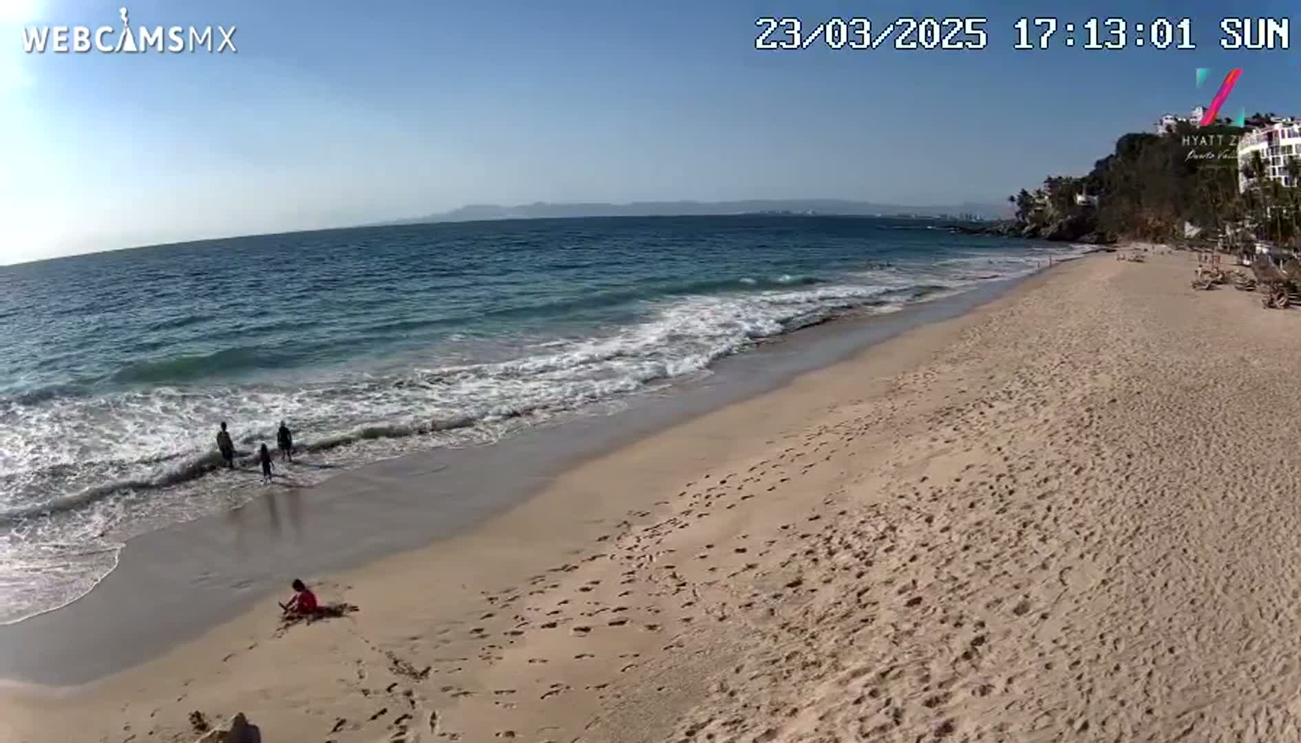 A sandy beach slopes down to a gently-lapping ocean under a clear blue sky, with a few people enjoying the water and shoreline.