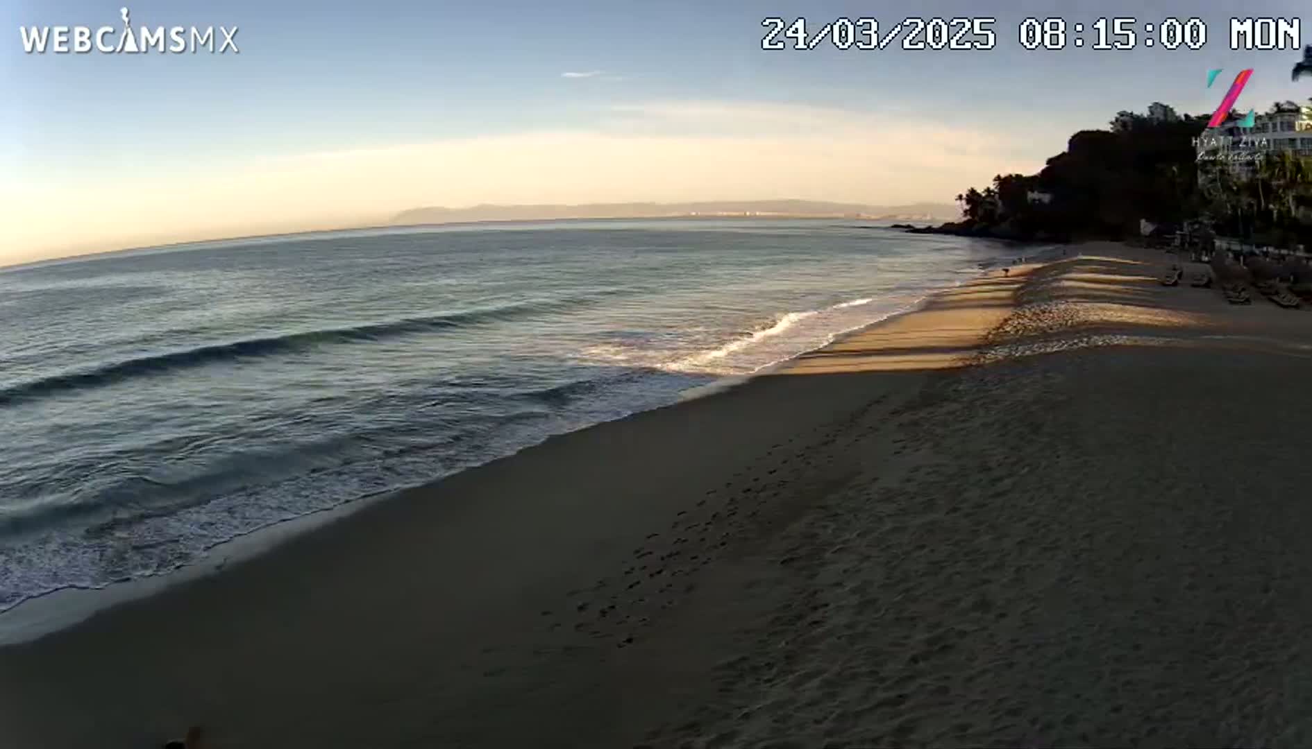 A sandy beach with footprints leading to calm ocean waves under a clear morning sky.