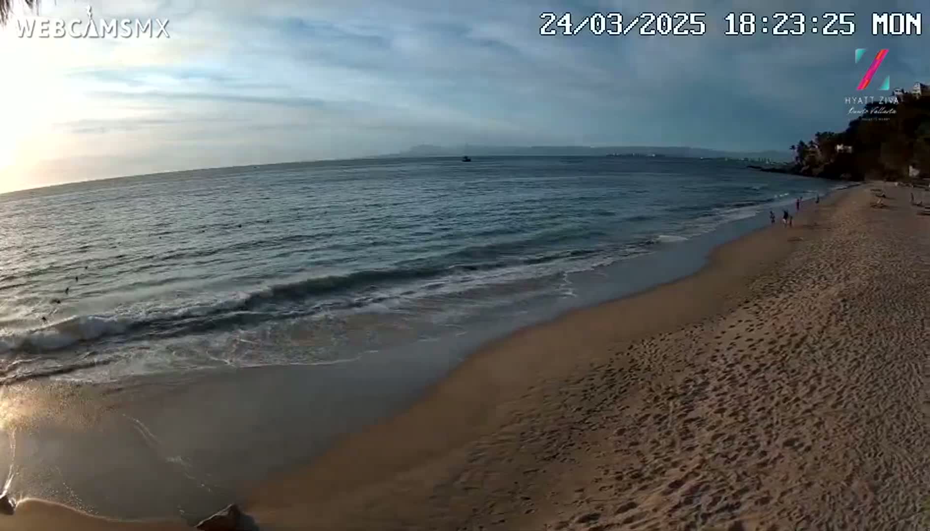 A sandy beach with gentle waves and a few people walking along the shoreline under a partly cloudy evening sky.