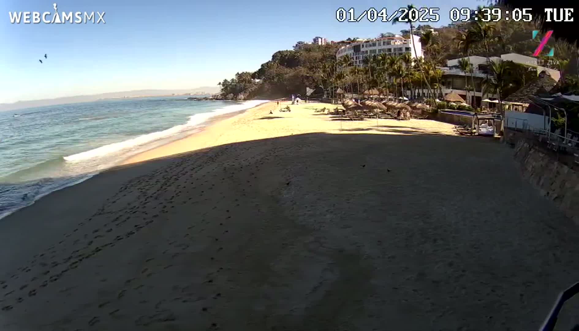 A sandy beach with footprints stretches from a calm ocean to a resort under a clear, sunny sky.