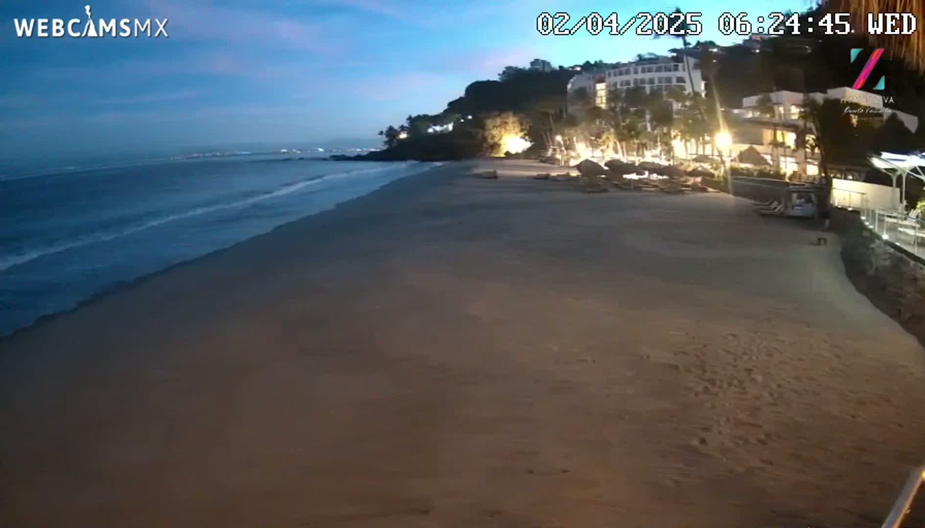 A sandy beach at twilight, with gentle waves lapping the shore, is bordered by a resort with illuminated buildings and palm trees.