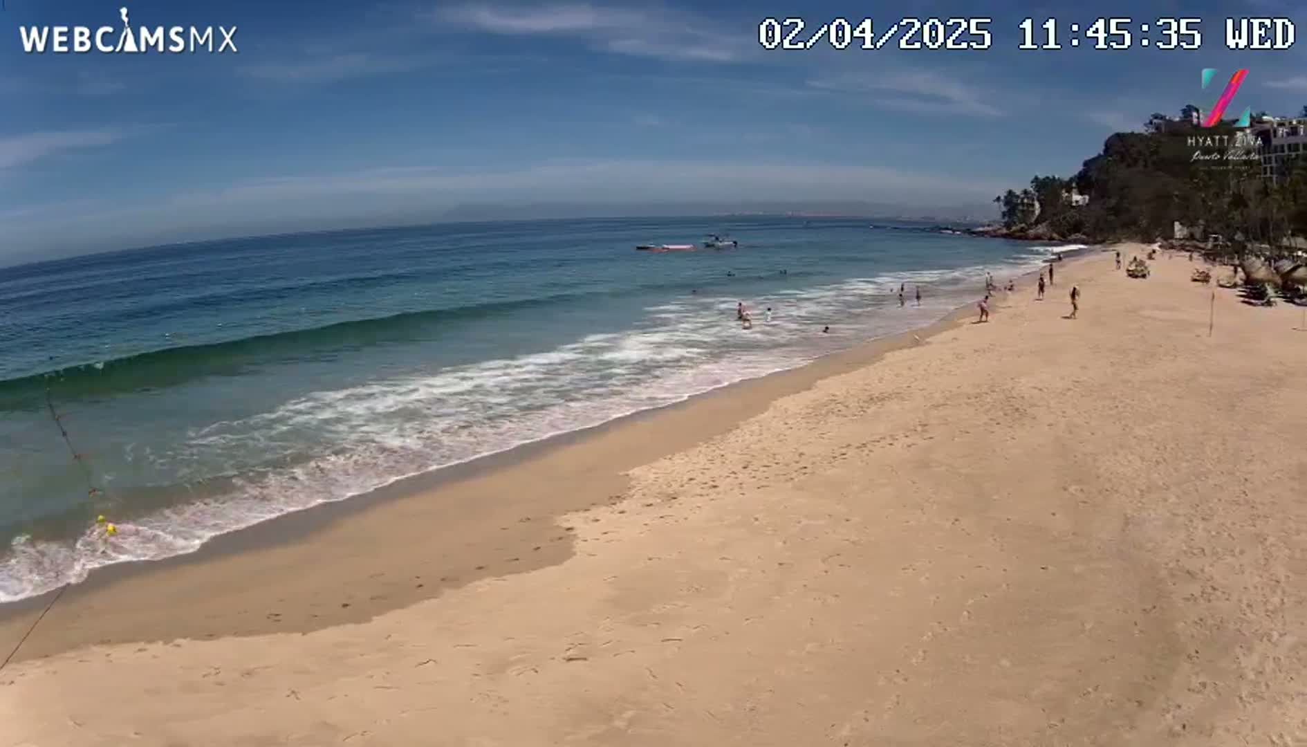 A sandy beach with a few people on it is gently lapped by ocean waves under a mostly sunny sky.