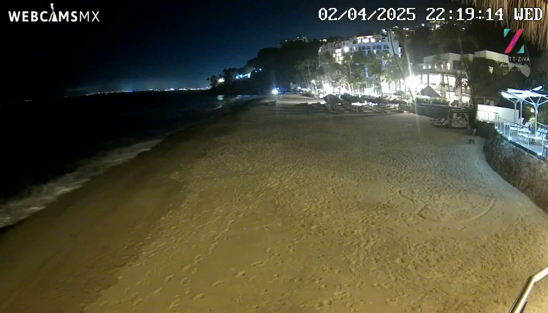 A sandy beach at night, gently lapped by waves, is adjacent to a brightly lit resort nestled against a dark hillside.