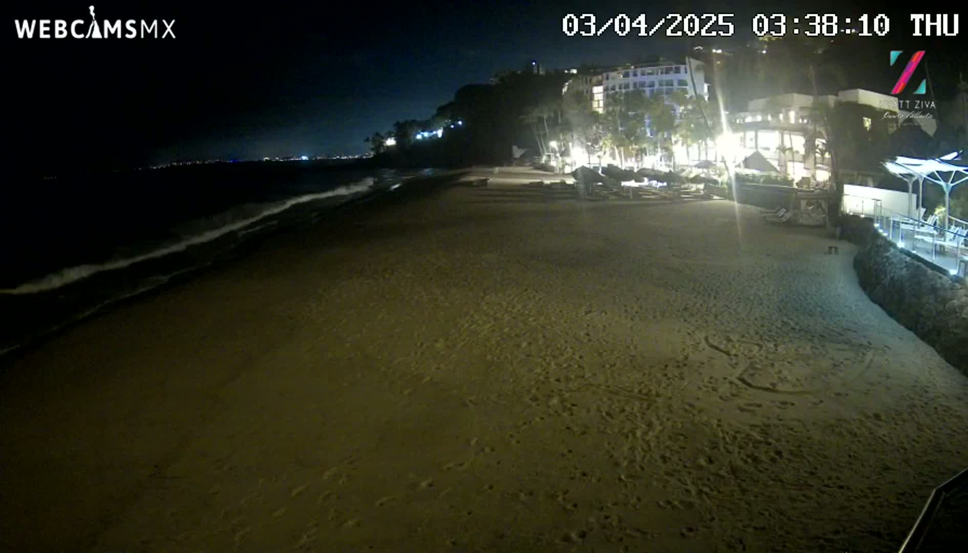 A dark sandy beach at night with gentle waves lapping the shore, and illuminated buildings on a cliff in the background.