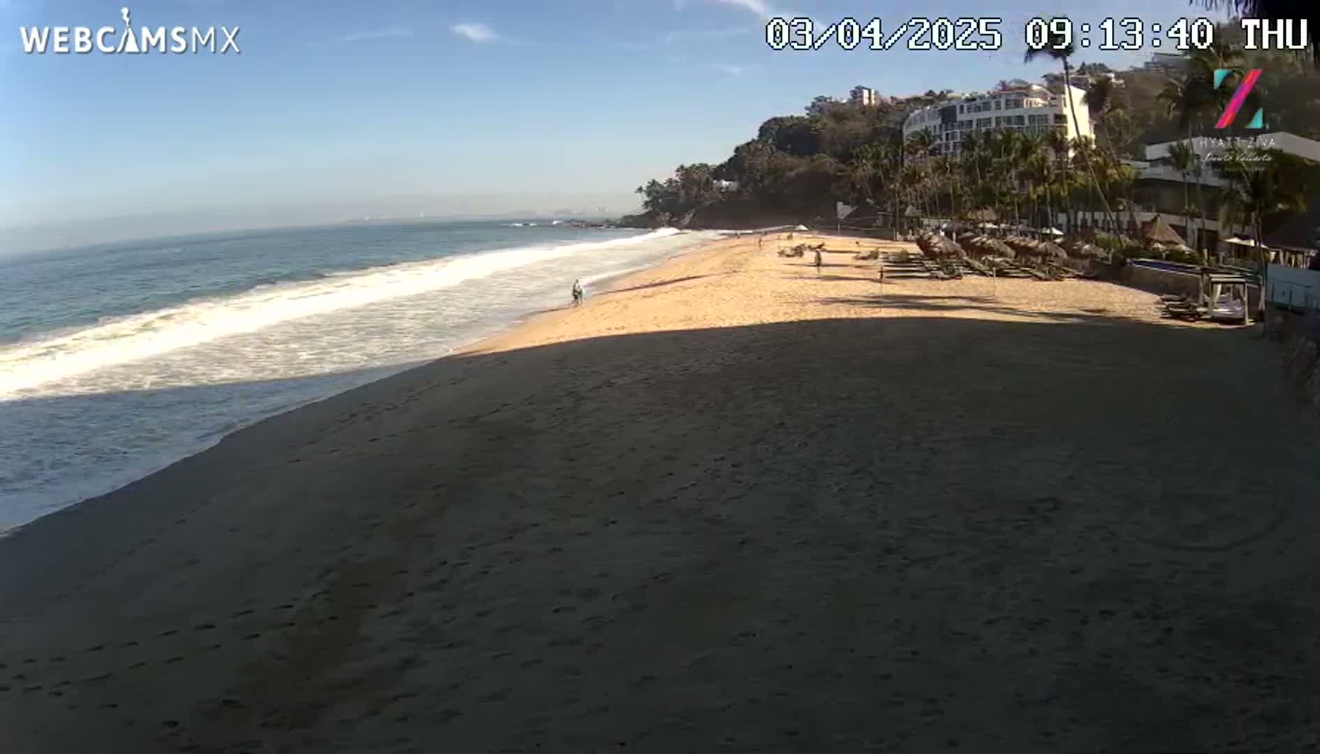 A sandy beach with gentle waves under a clear blue sky, showing a resort in the background.