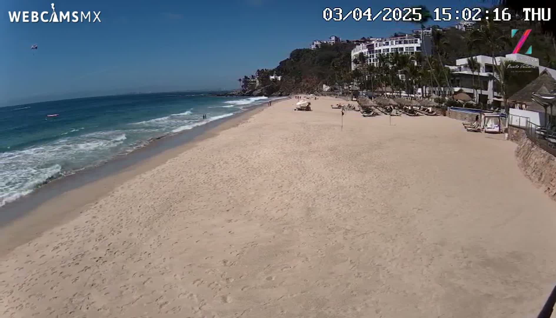 A sandy beach with light waves and a few people stretches out from a resort area under a clear blue sky.