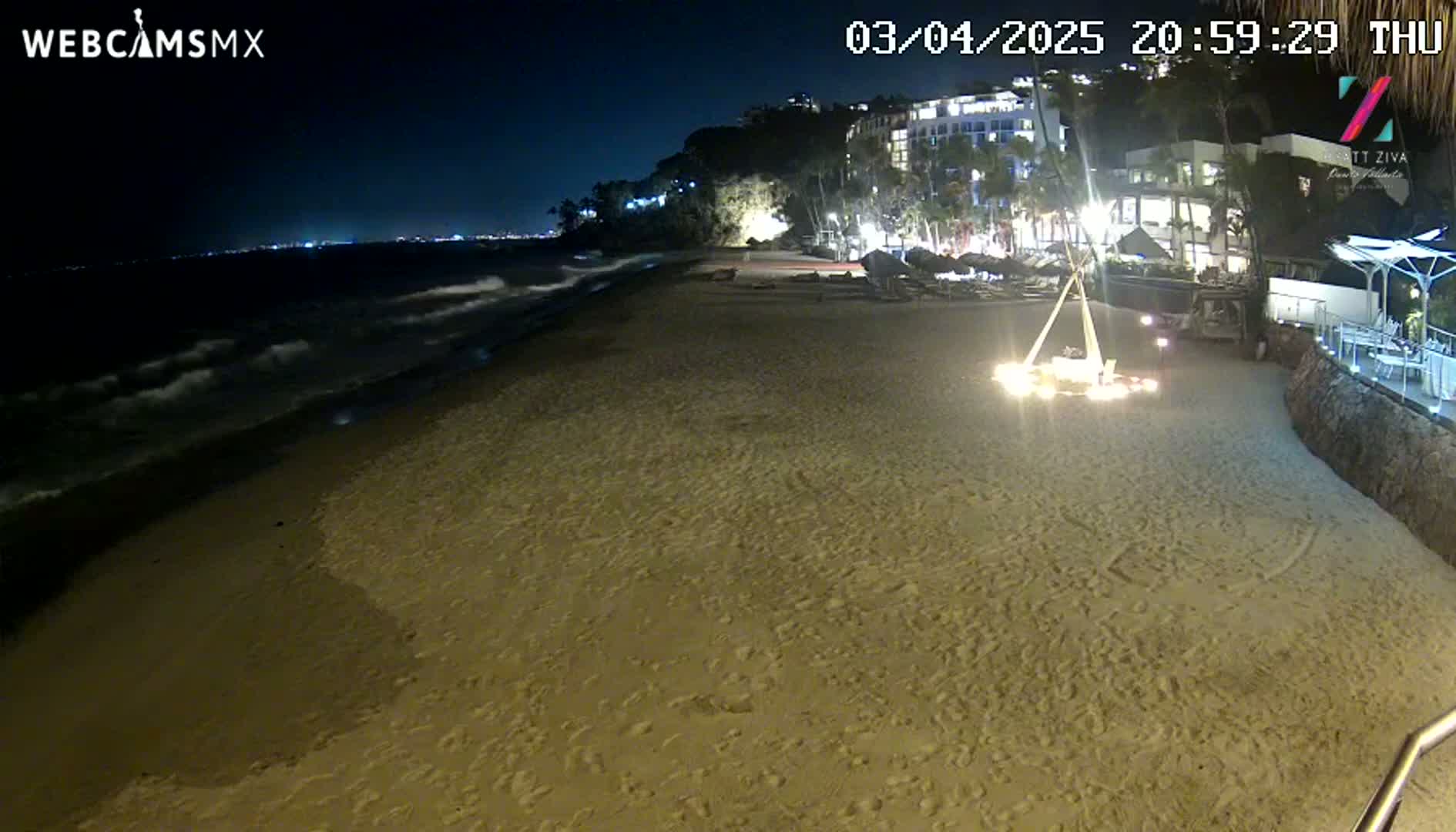 A sandy beach at night is illuminated by lights from buildings on a cliff overlooking the ocean, with gentle waves lapping the shore.