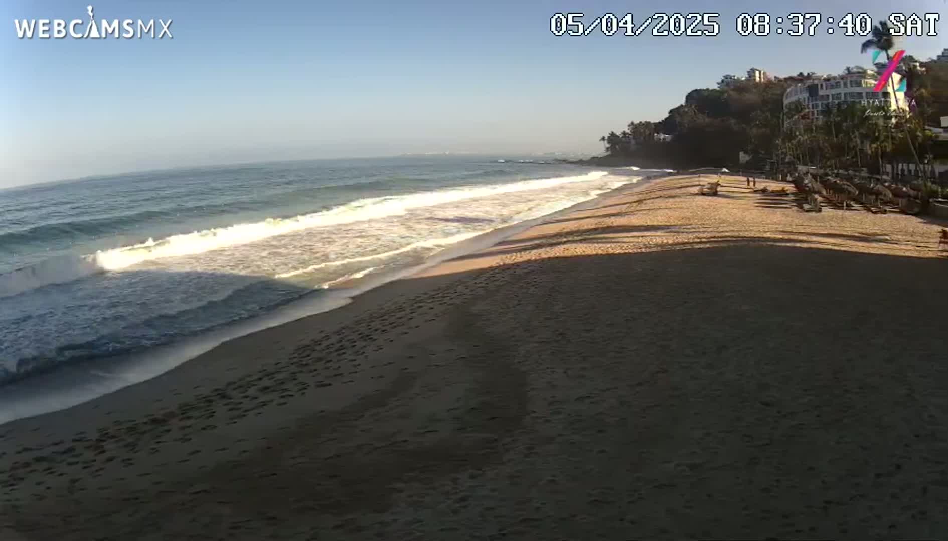 A sandy beach with gentle waves under a clear blue sky is visible, with a resort and vegetation in the background.