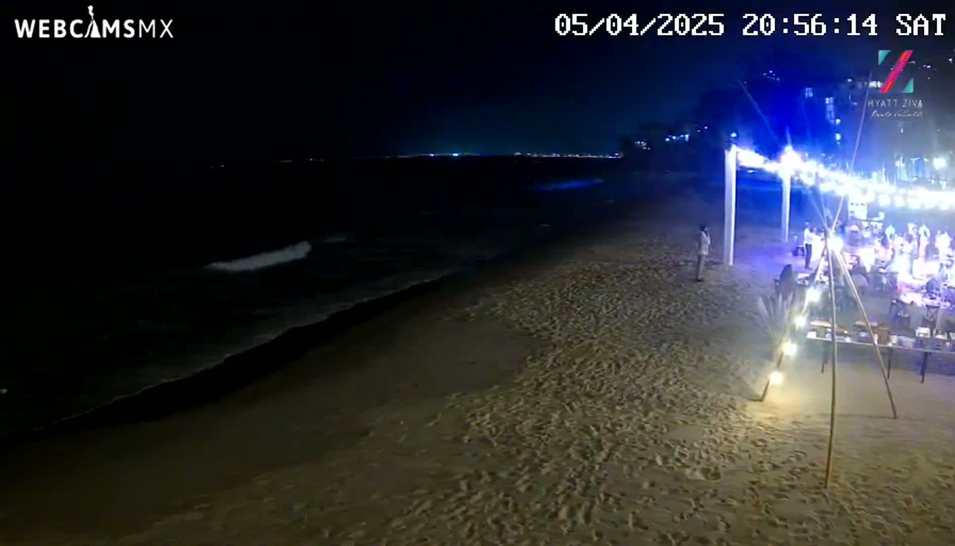 At night, gentle waves lap a sandy beach where a group of people are gathered near a lighted outdoor structure.