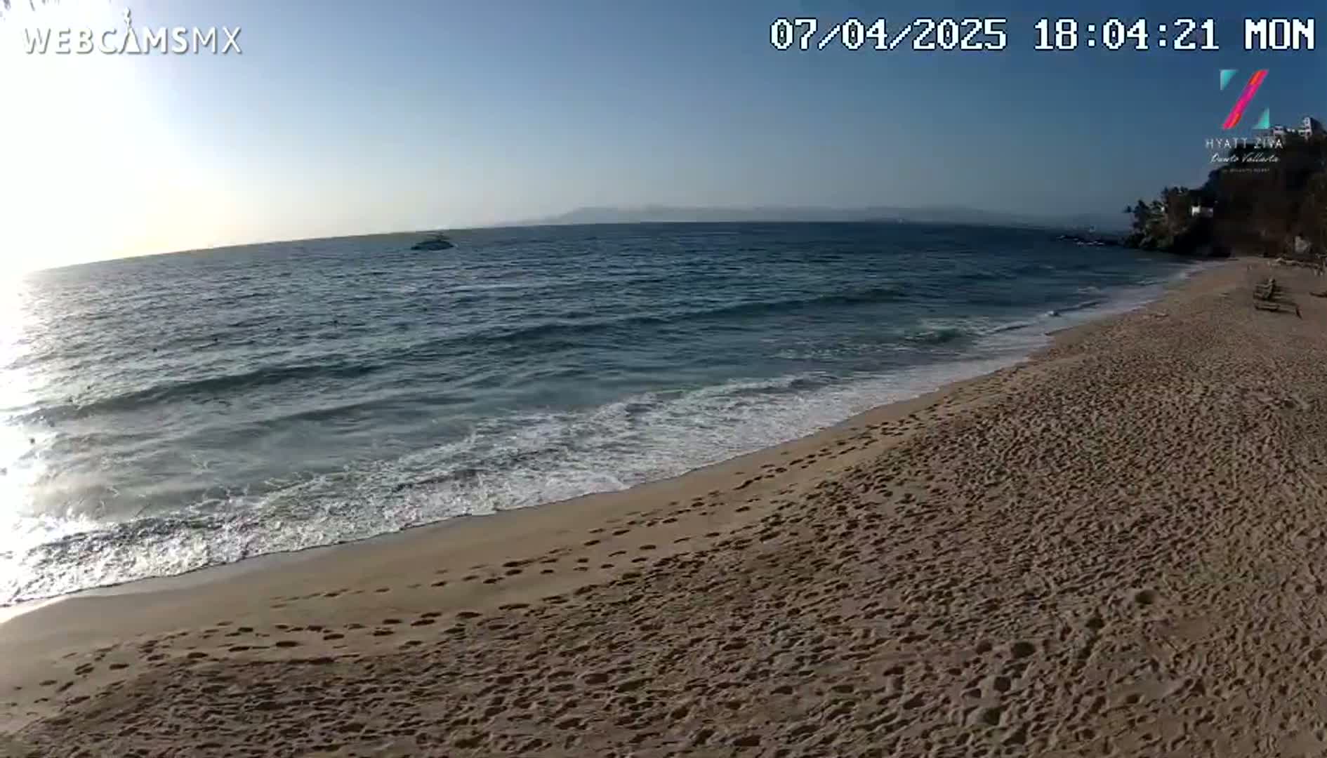 A sandy beach with many footprints stretches from a gently lapping ocean under a clear, sunny sky.