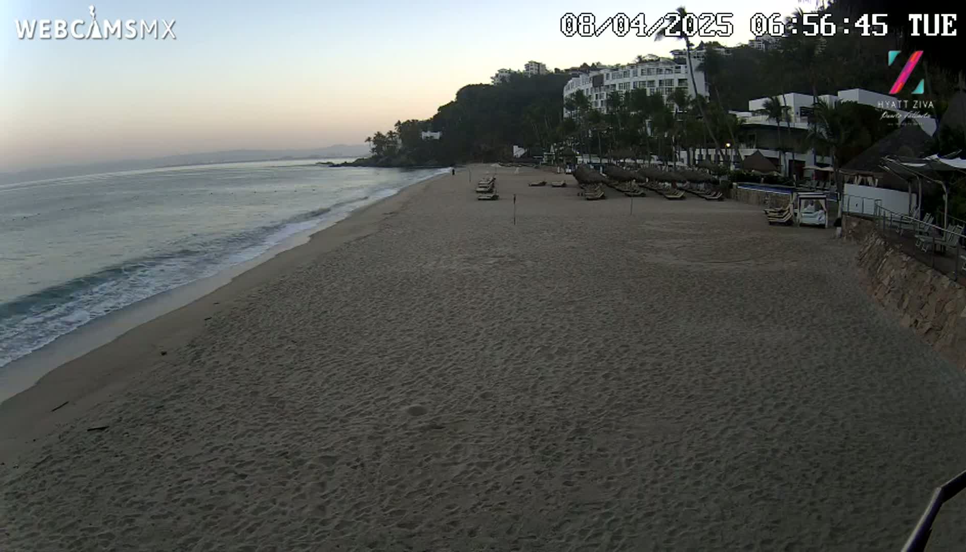 A sandy beach with calm waves and several lounge chairs in front of a resort under a twilight sky.