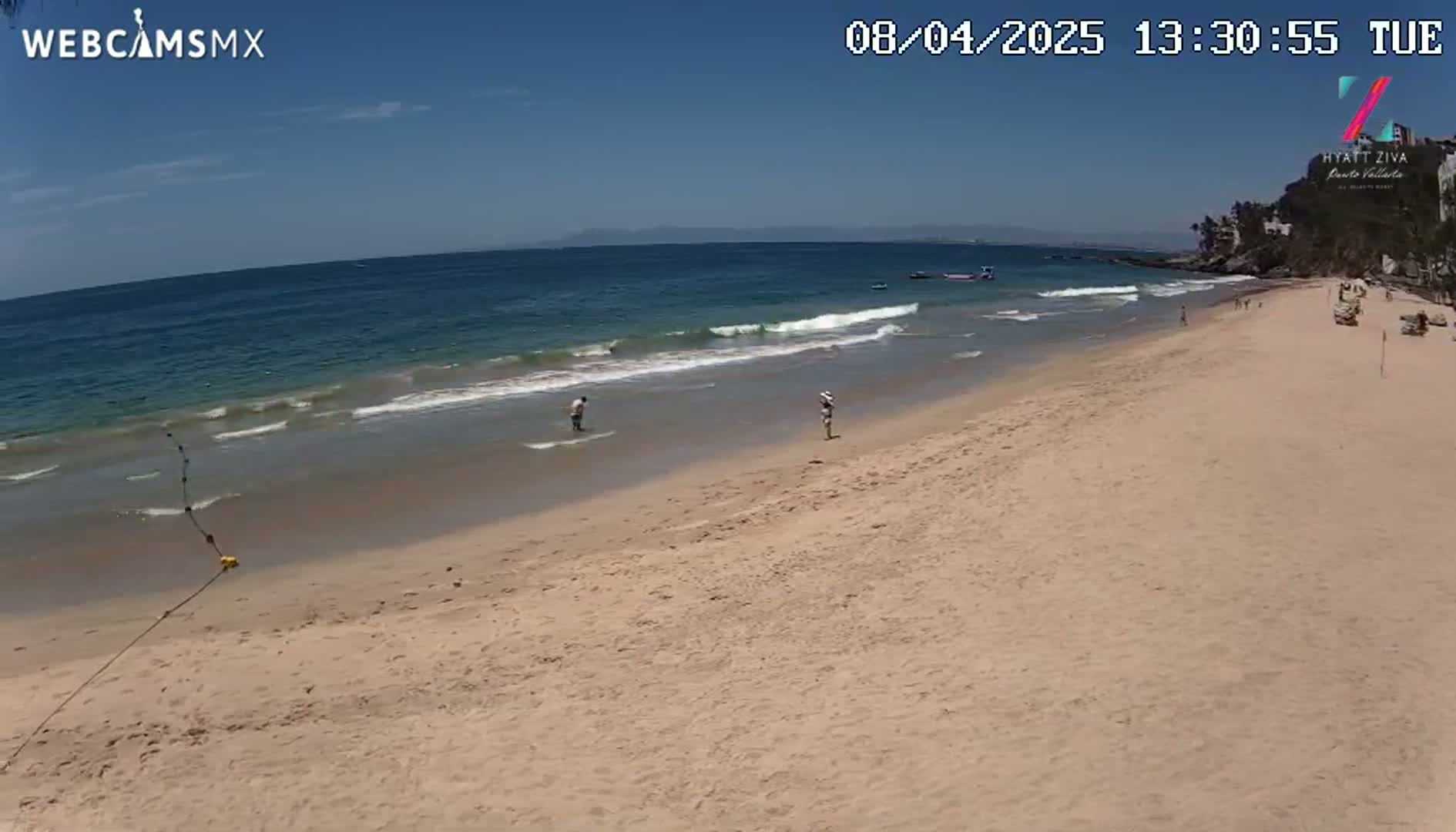 A sandy beach with calm ocean waves under a clear, sunny sky, and a few people are seen in the water and walking on the shore.
