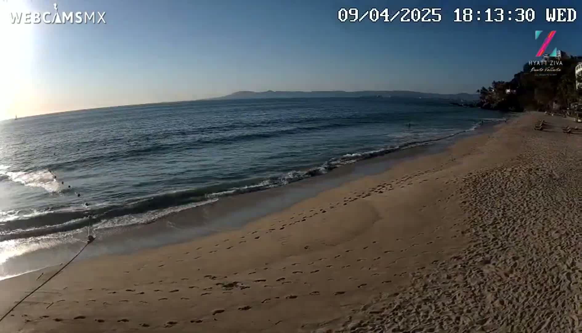 A sandy beach with footprints slopes gently down to a calm ocean under a clear, sunny sky.