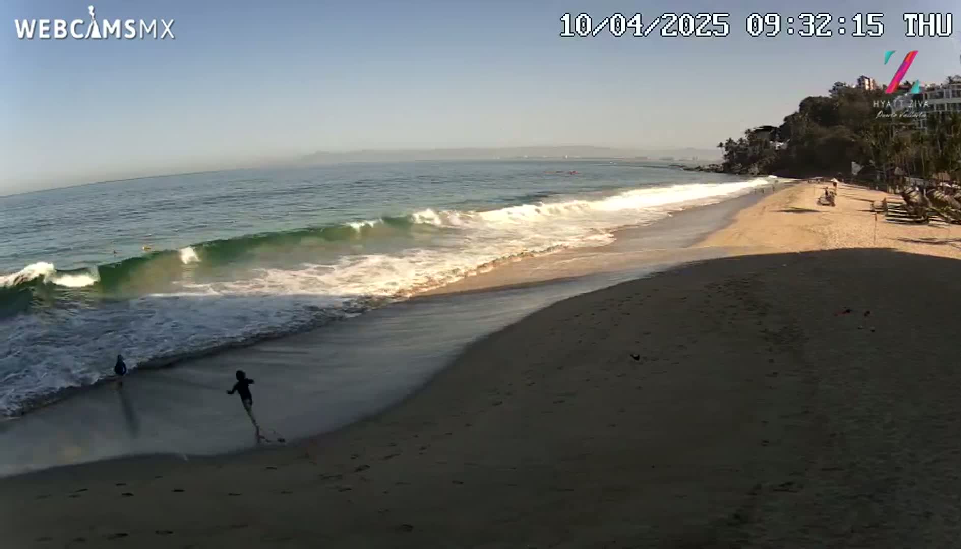 A sandy beach with a few people on it is gently lapped by waves under a clear, sunny sky.