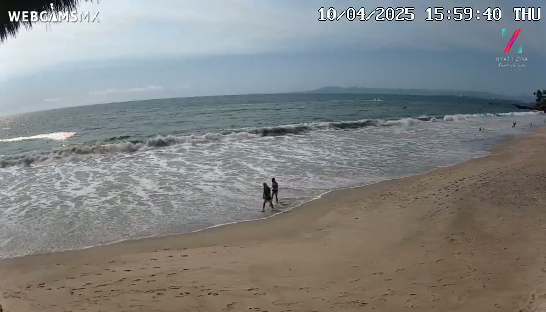 Two people walk along a sandy beach where small waves are rolling onto the shore under a mostly cloudy sky.