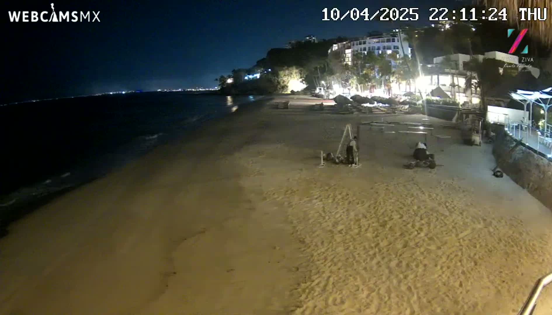 A mostly empty sandy beach at night is illuminated by lights from buildings on a cliff overlooking a calm sea.