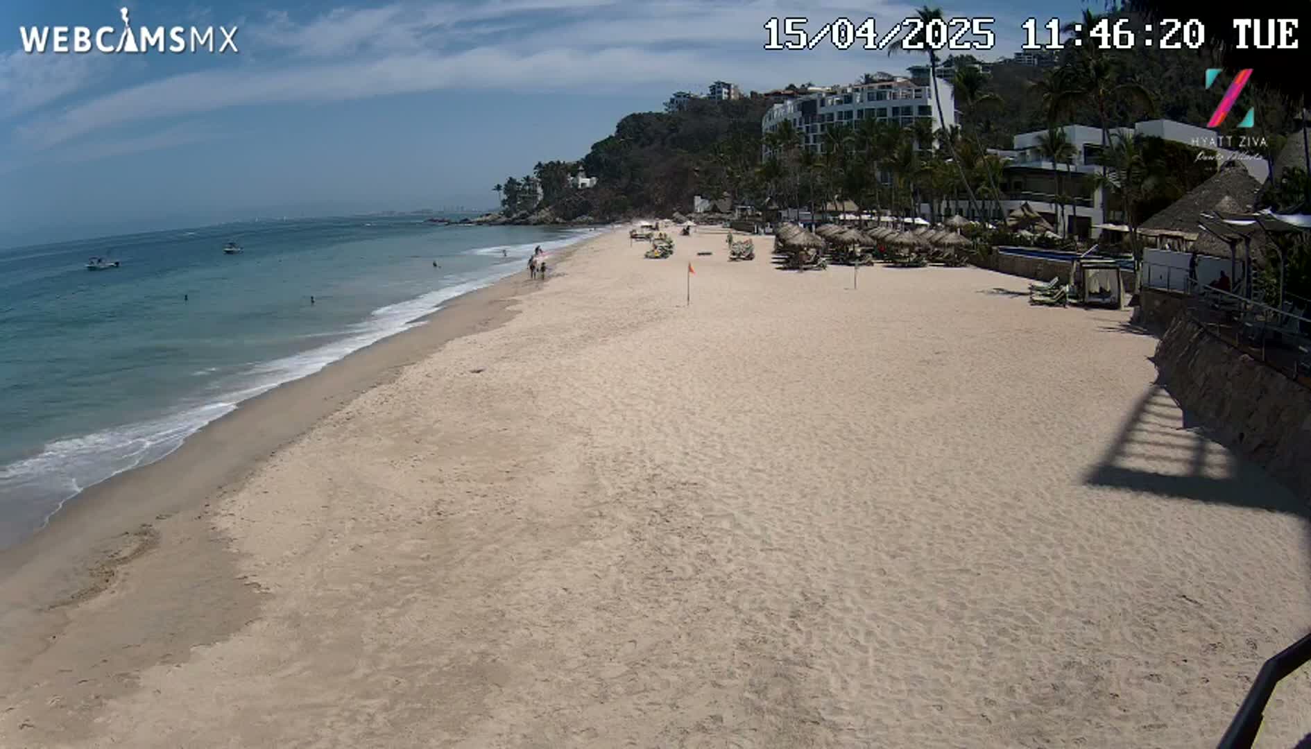 A sandy beach with calm ocean waves stretches from a resort with palm trees under a mostly sunny sky.