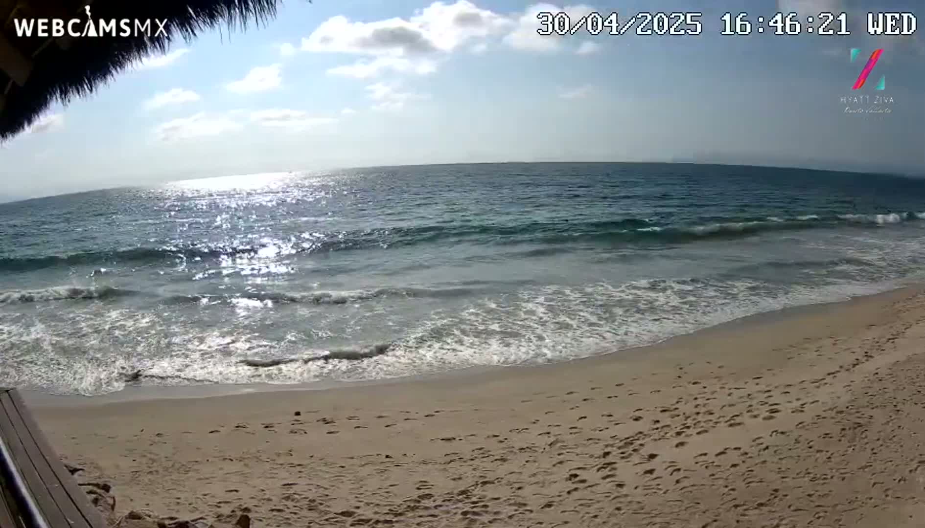 A sandy beach with footprints is shown, where small waves from a sparkling ocean under a partly cloudy sky lap the shore.