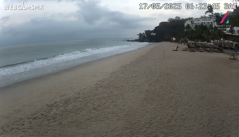 A sandy beach with gentle waves under an overcast sky, showing a resort in the background.