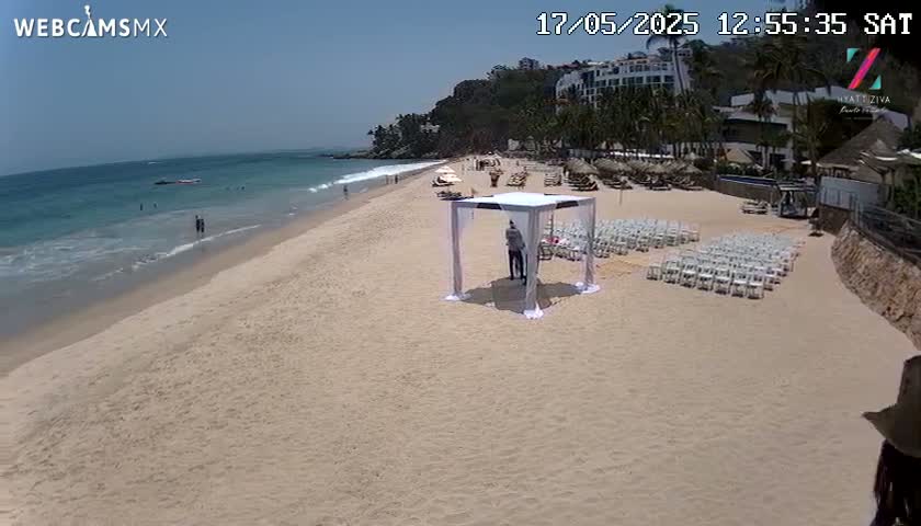 A sandy beach with calm ocean water under a sunny sky shows a wedding ceremony setup with white chairs and a canopy.