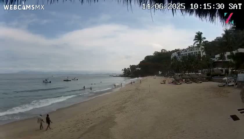 A sandy beach with calm ocean waves, a few people, and several small boats in the distance under a cloudy sky.