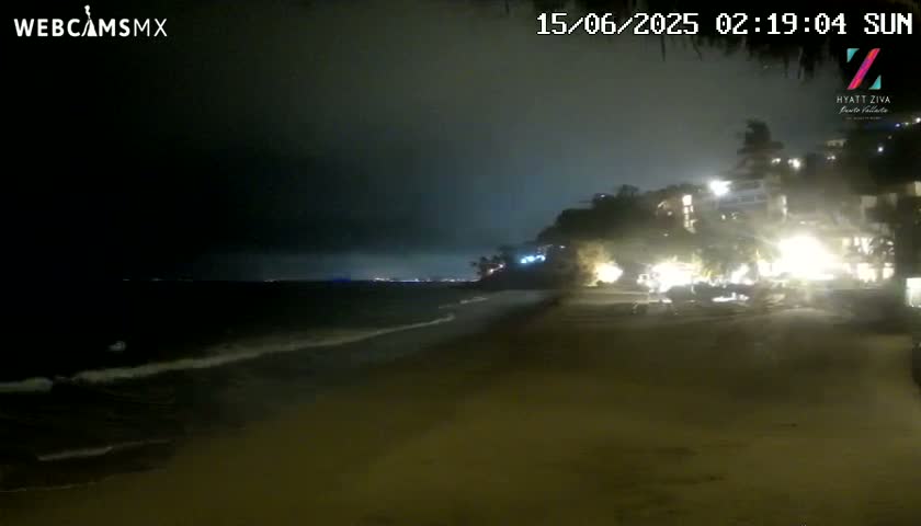 A nighttime beach scene shows dark, gentle waves lapping the shore under a mostly cloudy sky, with brightly lit buildings on a cliff in the background.