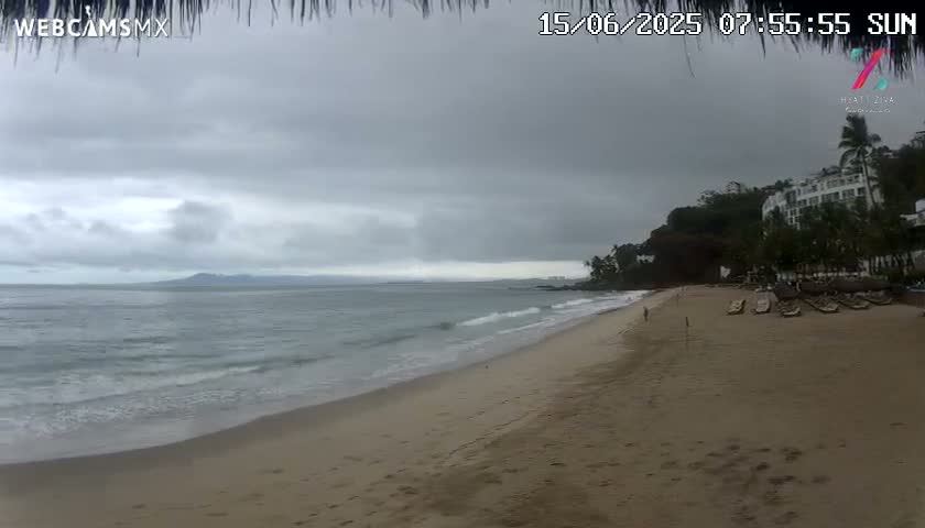 A sandy beach with gentle waves laps the shore under an overcast sky, with a large resort visible in the background.