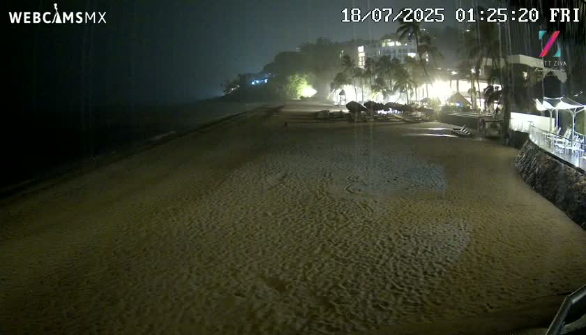 A dark, sandy beach at night is shown under rainy conditions, with a resort and palm trees visible in the background.
