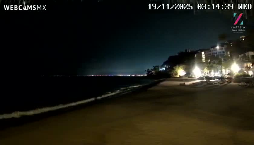 A clear night reveals a dark beach with gentle waves lapping the shore, illuminated resort buildings on the right, and distant city lights twinkling across the water.