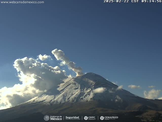 A snow-capped volcano erupts a plume of ash into a partly cloudy sky.