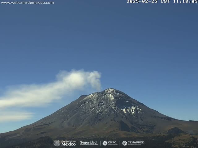 A snow-capped volcano emits a plume of white smoke under a clear blue sky.