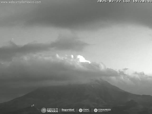 A grayscale image shows a mostly cloudy sky over a large, dark mountain at dusk or dawn.