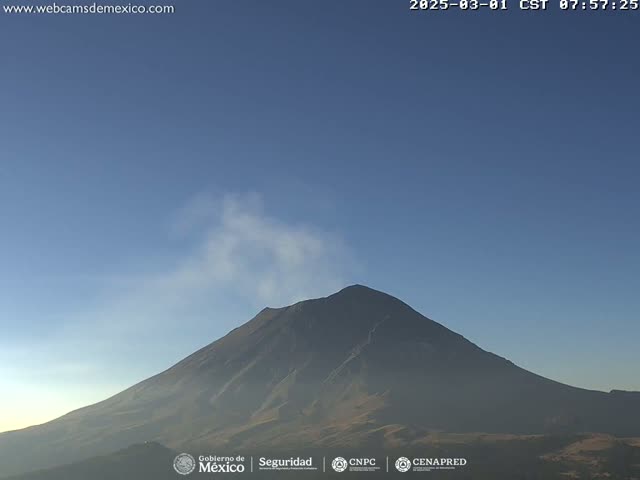 A large, dark-colored mountain with a plume of light-colored smoke rising from its peak is visible under a clear, light-blue sky.