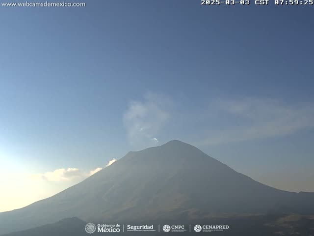 A large, dark-colored volcano with a plume of smoke or steam rising from its peak is visible under a mostly clear, light-blue sky.